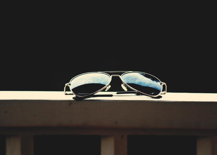 Un gros plan de lunettes de soleil aviateur reflétant le ciel, sur fond sombre.