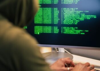 Personne portant un hoodie tapant du code sur un écran d'ordinateur avec du texte vert, représentant une scène de hacking.