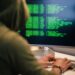 Personne portant un hoodie tapant du code sur un écran d'ordinateur avec du texte vert, représentant une scène de hacking.