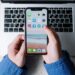 Vue rapprochée de mains tenant un smartphone avec des applications sur un clavier d'ordinateur portable.