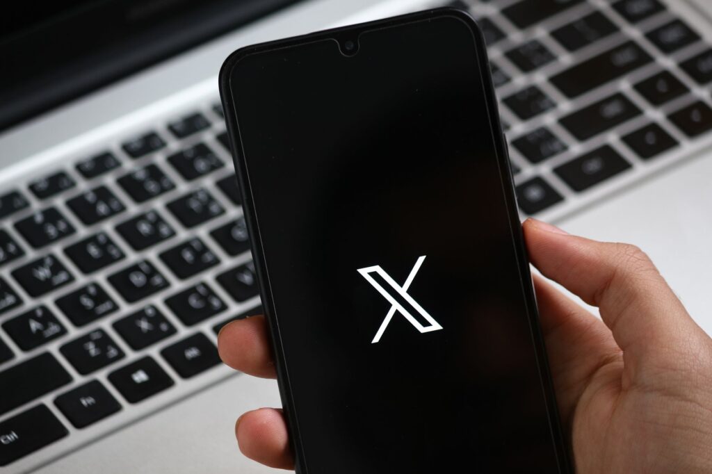 Une main tient un smartphone affichant une interface d'application moderne, avec un clavier d'ordinateur portable en arrière-plan.