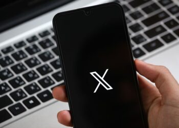 Une main tient un smartphone affichant une interface d'application moderne, avec un clavier d'ordinateur portable en arrière-plan.