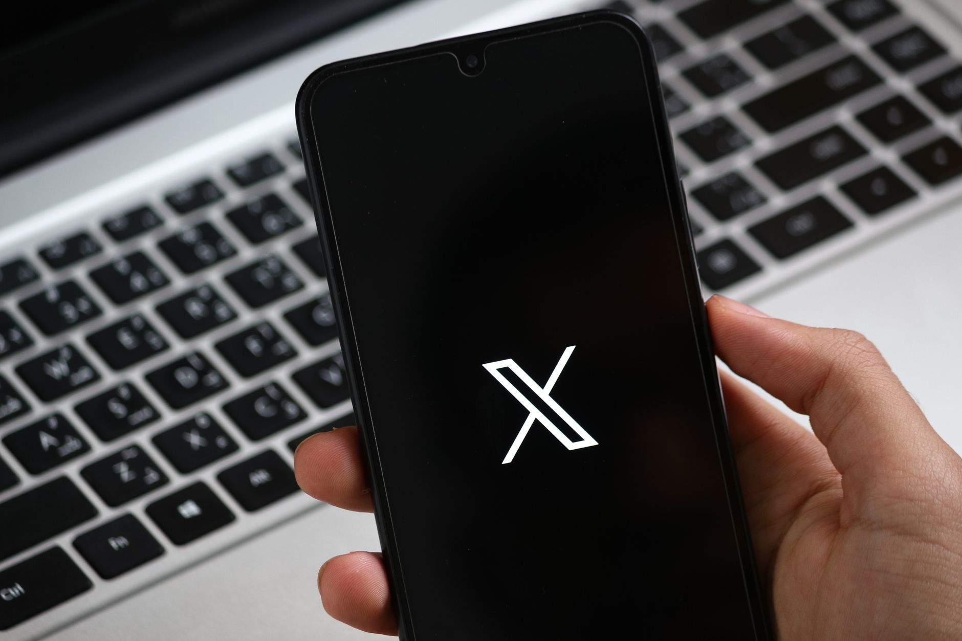 Une main tient un smartphone affichant une interface d'application moderne, avec un clavier d'ordinateur portable en arrière-plan.